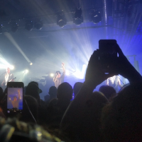 A band performing on a stage with bright lights in a dark room full of a large crowd.